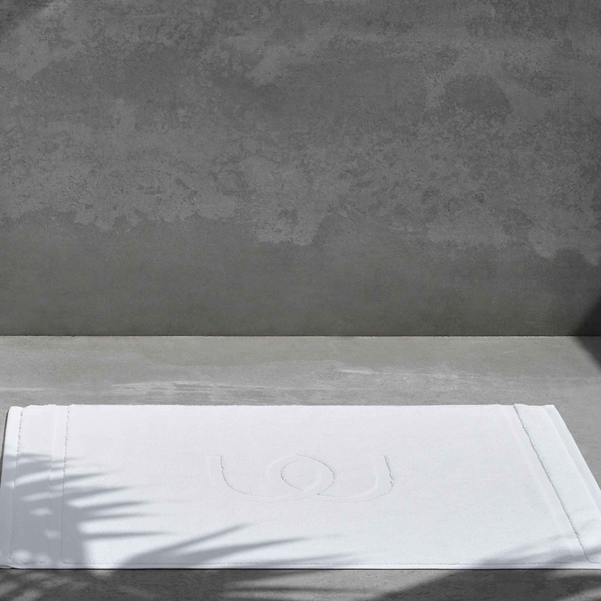 White bath mat with jacquard logo in the centre on a concrete floor with subtle palm shadow